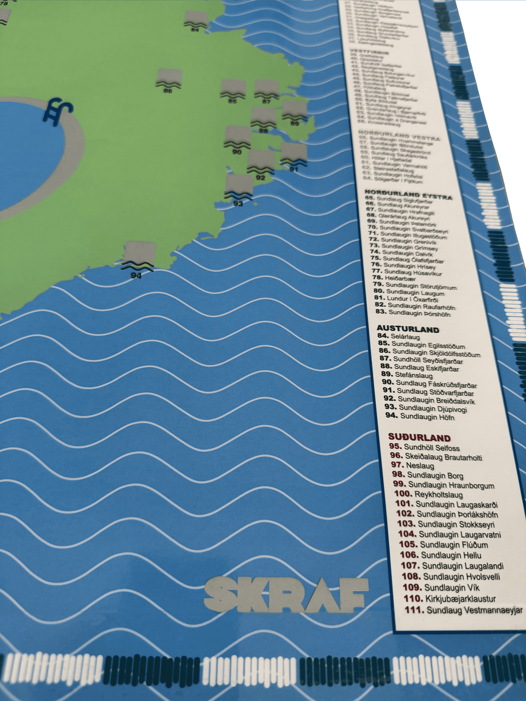 Nærmynd af Skraf skafkorti fyrir sundlaug Íslands sem sýnir skafreiti á Austurlandi og yfirlitslista yfir allar sundlaugar landsins flokkaða eftir landshlutum með númerum og nöfnum.